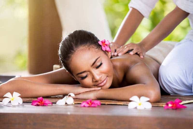 A woman with a pink flower in her hair lies on her stomach, eyes closed and smiling, enjoying an outdoor massage. Colorful flowers are scattered around her on a wooden surface, with a lush green background completing the peaceful scene.