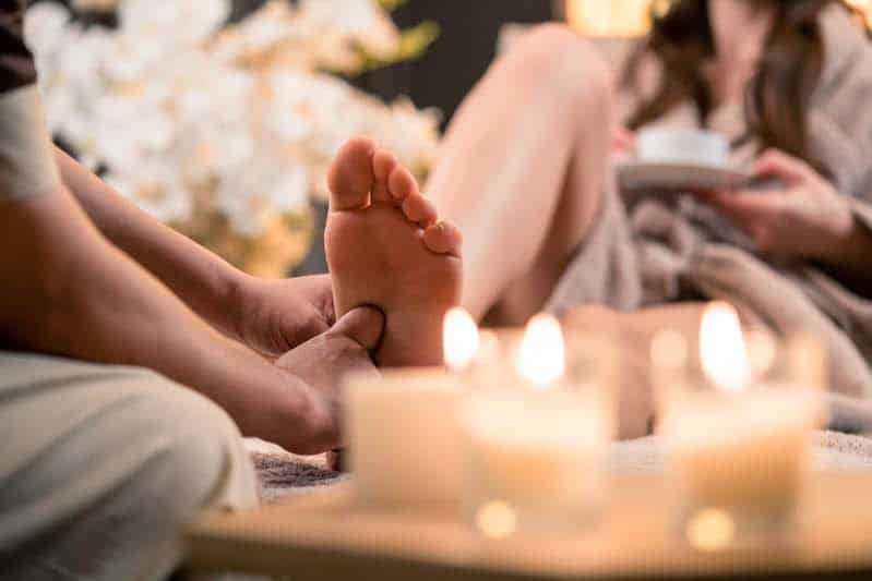 A person enjoys a relaxing reflexology foot massage in a cozy setting with lit candles, while another person in a robe sips from a cup and sits nearby.