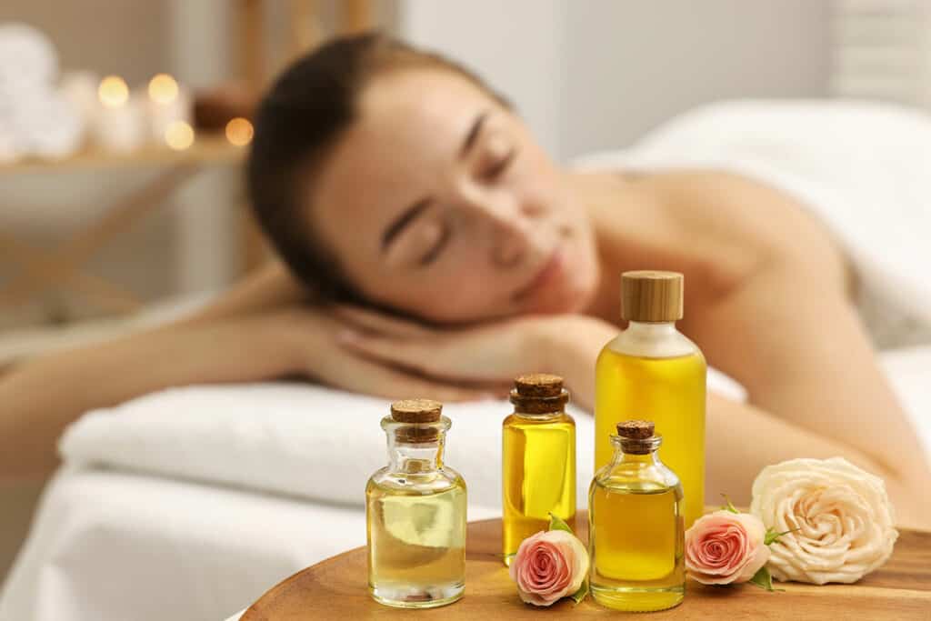 A woman relaxes with her eyes closed on a massage table, with bottles of essential oils and pink roses in the foreground, creating a calm spa atmosphere.