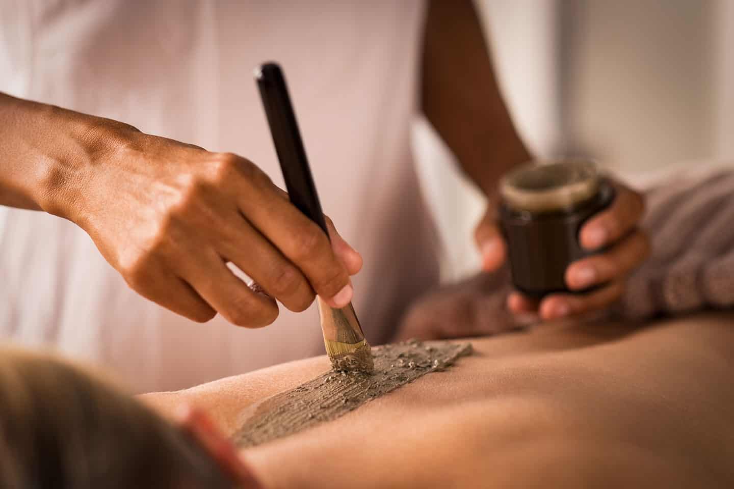 A person applies a green mud or clay mask with a brush to someone’s bare back, likely during a spa treatment. A small jar with more of the substance is held in the other hand.