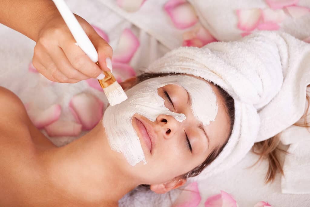 A person with a towel wrapped around their head lies with eyes closed as a luxurious facial mask is applied with a brush; pink rose petals and towels surround them in a serene spa setting.
