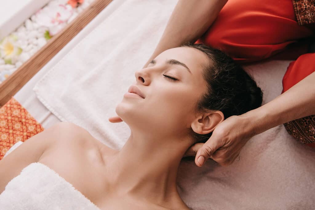Woman lying on a massage table with eyes closed, wrapped in a white towel, receiving a relaxing neck massage from a therapist dressed in red clothing.