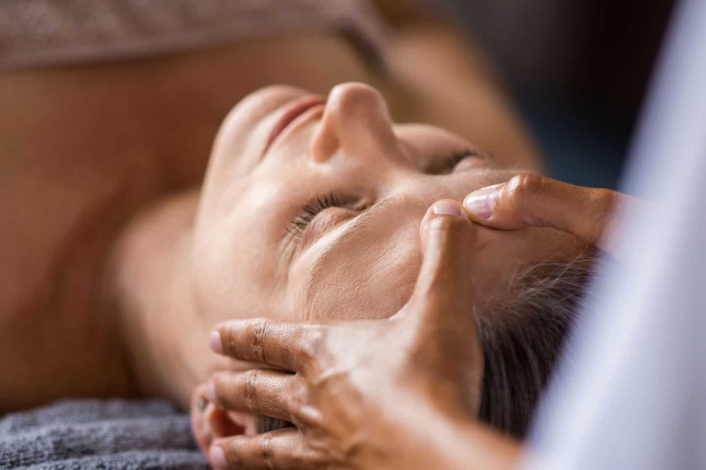 A person lies with eyes closed while another person gently massages their forehead, suggesting relaxation or a spa treatment.