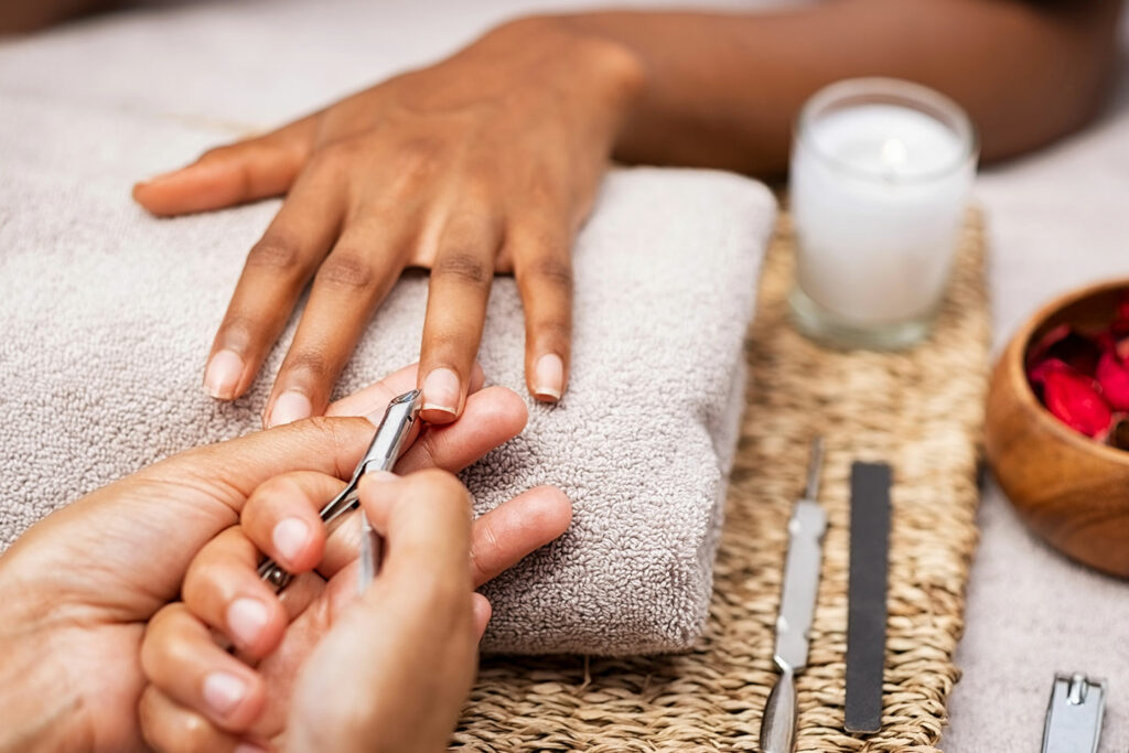 A close-up of a person receiving a manicure; one hand rests on a towel while the other hand uses a cuticle tool. Nearby are a candle, nail care tools, and a bowl of rose petals on a woven mat.