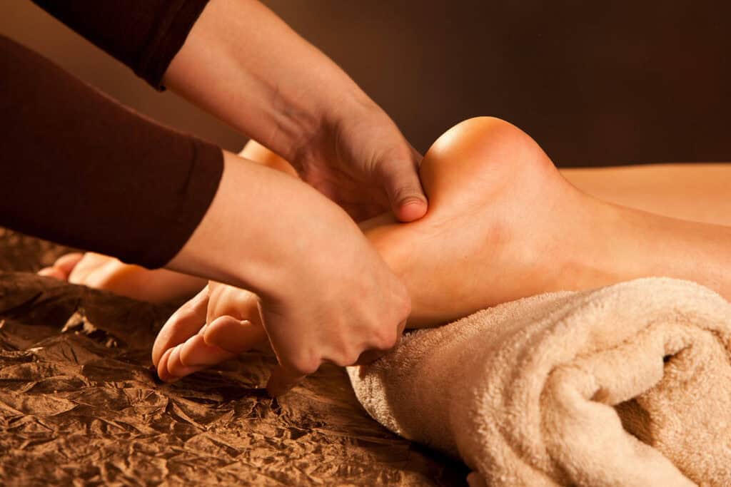 A person receives a relaxing reflexology foot massage, with their ankle resting on a rolled towel and hands gently pressing the heel and arch. The setting is warm and calming.