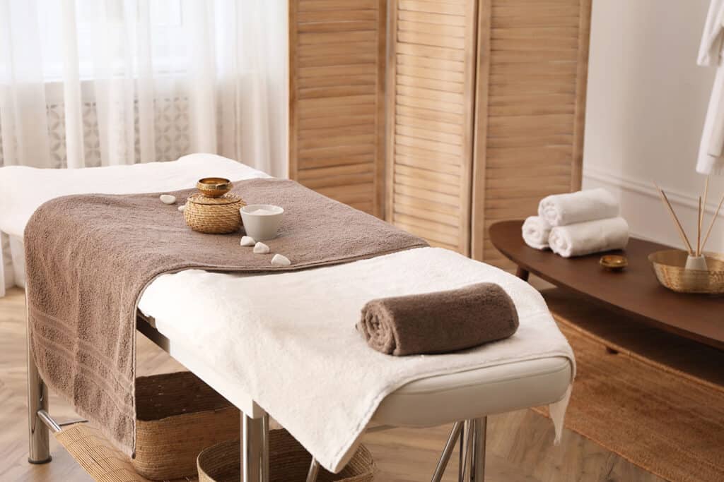 A spa room with a massage table covered in white and brown towels, small bowls, and stones. rolled towels, woven baskets, and reed diffusers are arranged nearby, with soft lighting and a wooden privacy screen in the background.
