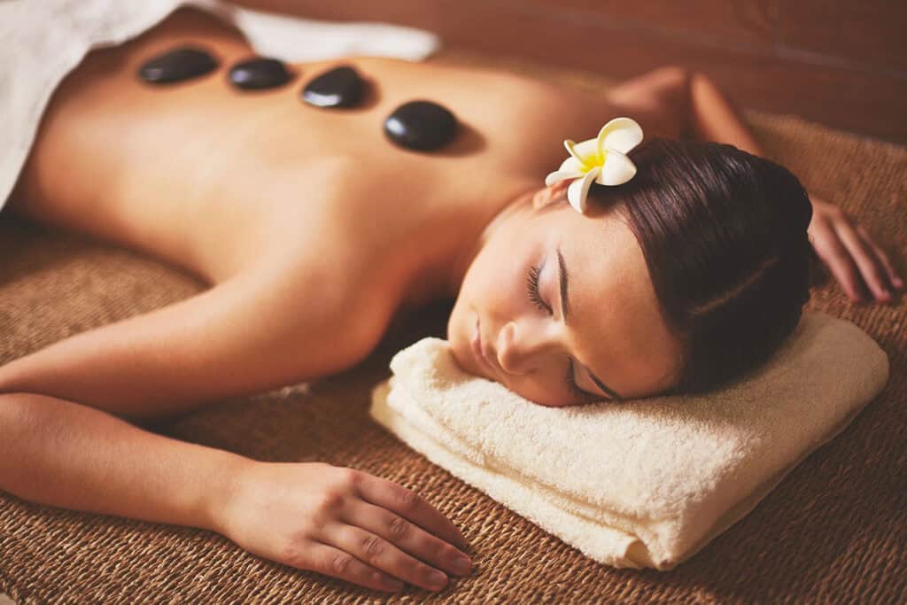 A woman with a flower in her hair lies face down on a towel, relaxing during a Bali Spa hot stone massage, with four black stones placed along her bare back.