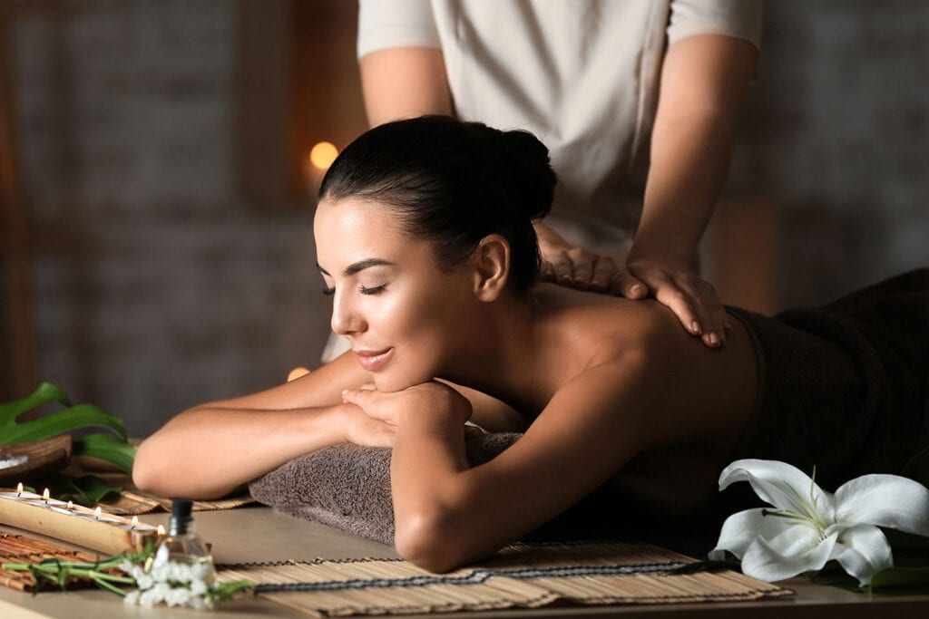 A woman with dark hair lies face down on a massage table at a Spa, receiving a relaxing back massage. Eyes closed, she looks serene amid elegant spa decor, including a white lily and softly glowing candles.