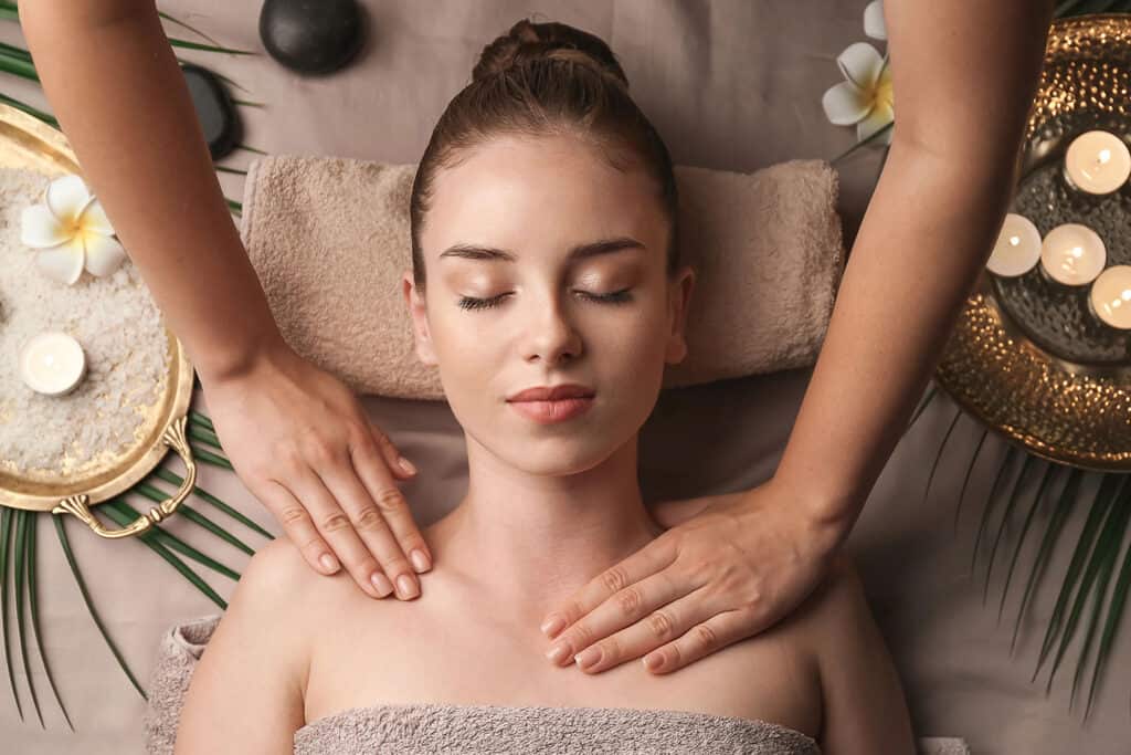 A woman with closed eyes receives a relaxing shoulder massage at a Spa, lying on a towel and surrounded by candles, flowers, and tranquil spa decor.
