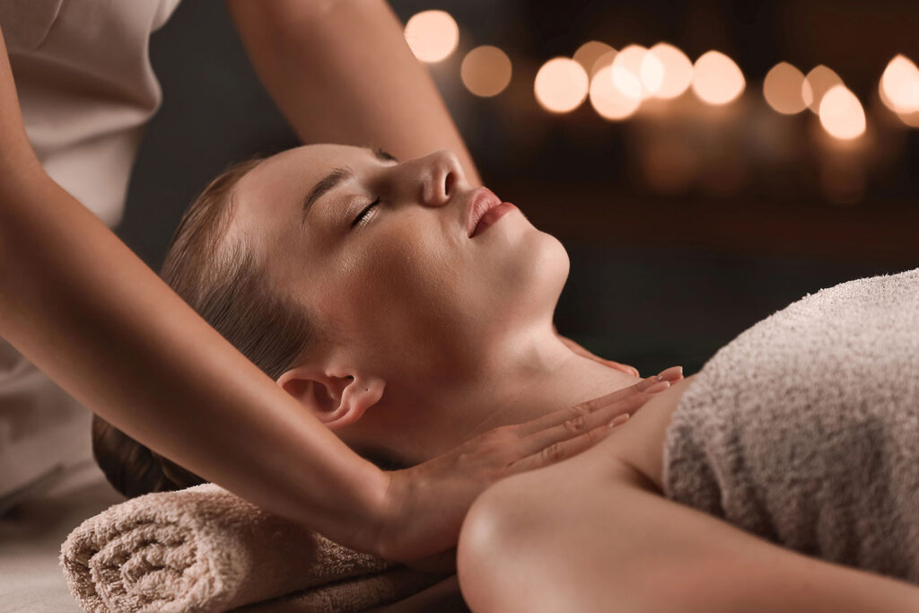 A woman with closed eyes relaxes on a towel-covered massage table while receiving a neck and shoulder massage in the softly lit, tranquil ambiance of a Bali Spa.