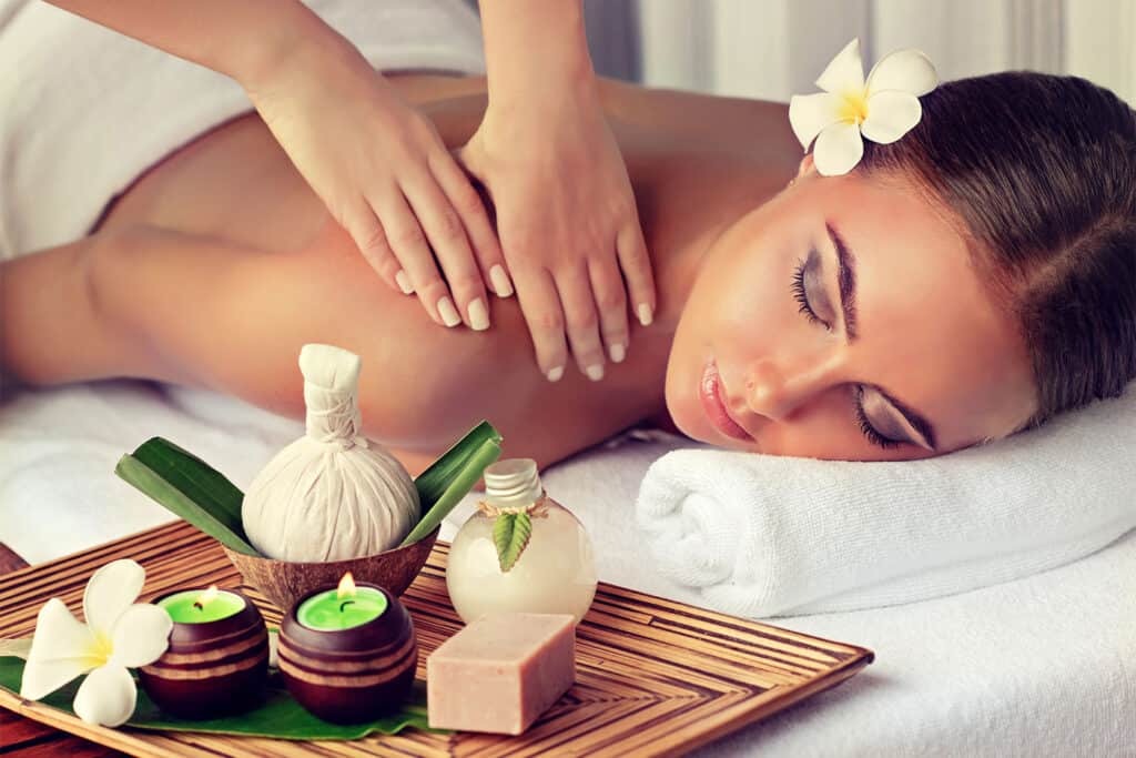 A woman with a flower in her hair receives a back massage at a spa. she is lying on a white towel, surrounded by candles, herbal compress, soap, and flowers on a wooden tray. the setting is calm and relaxing.