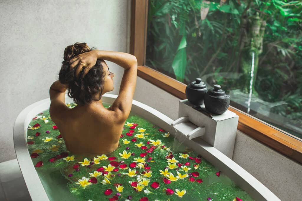 A person bathes in a tub filled with water and floating yellow and red flowers, facing a large window with a view of lush green plants outside. two black jars sit on the edge of the tub.
